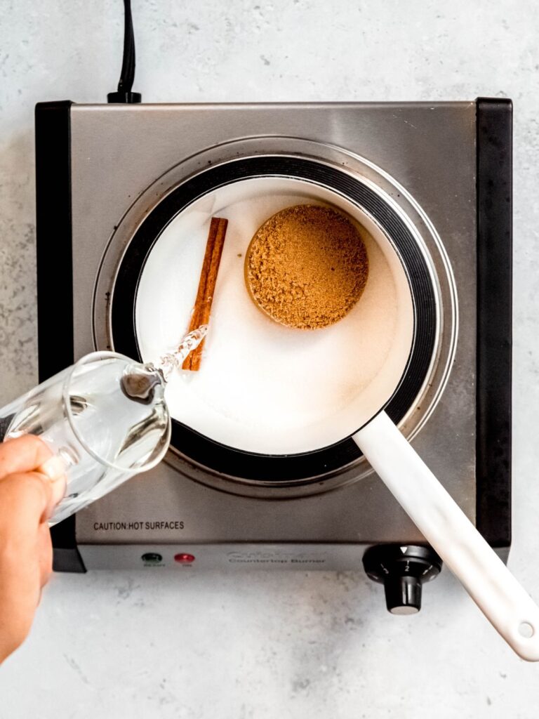 action shot of water being poured into a saucepan with a cinnamon stick, brown sugar and white sugar.