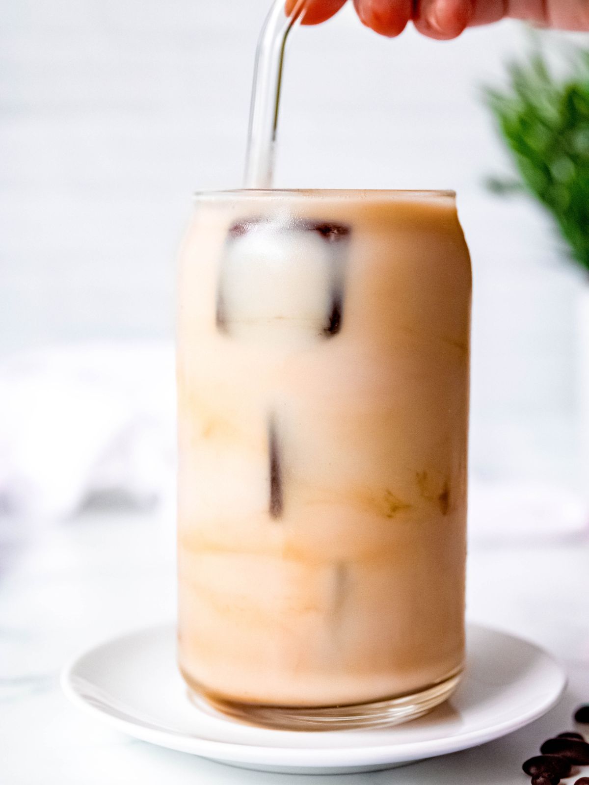 hand using a glass straw to swirl the coffee ice cubes in a glass of sweetened milk.