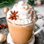 square hero image of an eggnog latte in a clear mug topped with whipped cream and a whole star anise.