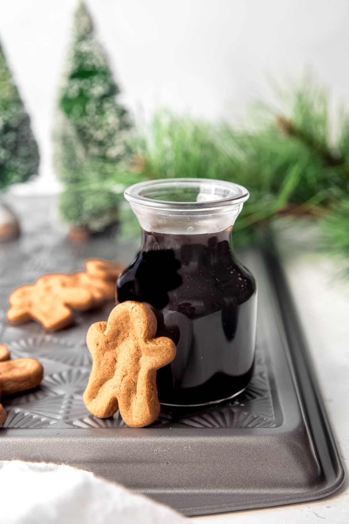 A small decanter of gingerbread syrup on a metal pan next to gingerbread man cookies.