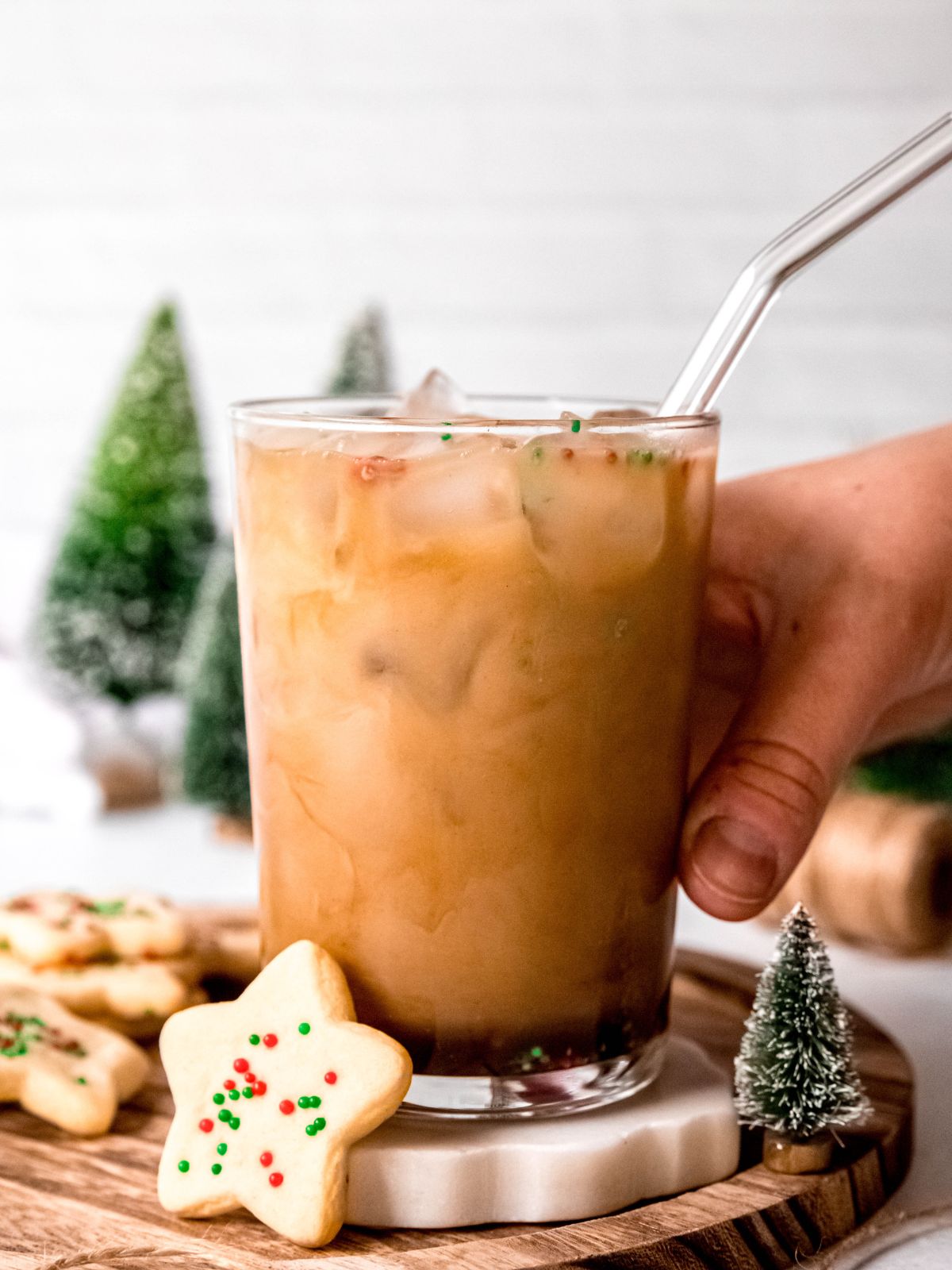 hand grabbing a clear glass filled with an iced sugar cookie latte with a glass straw. 