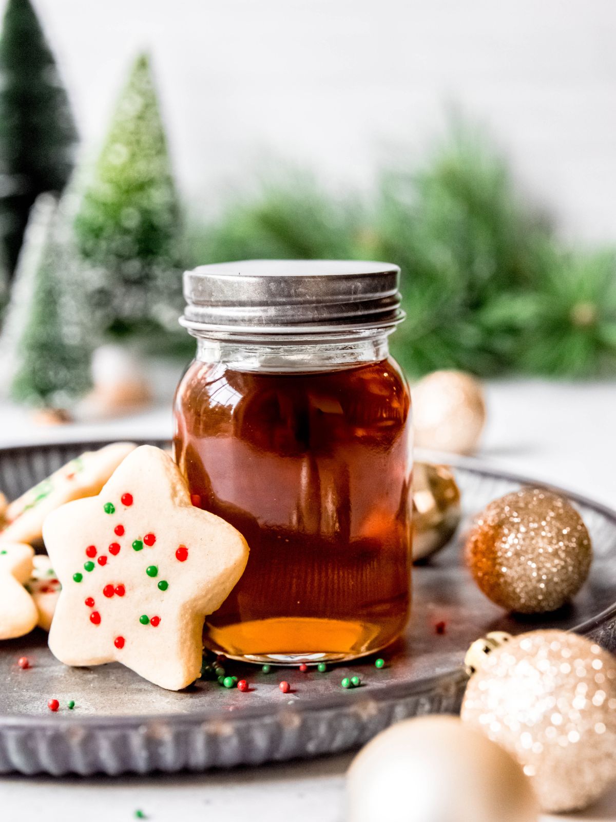 sugar cookie syrup in a small, silver lidded jar with star-shaped sugar cookies and small gold ornaments on a silver tray.