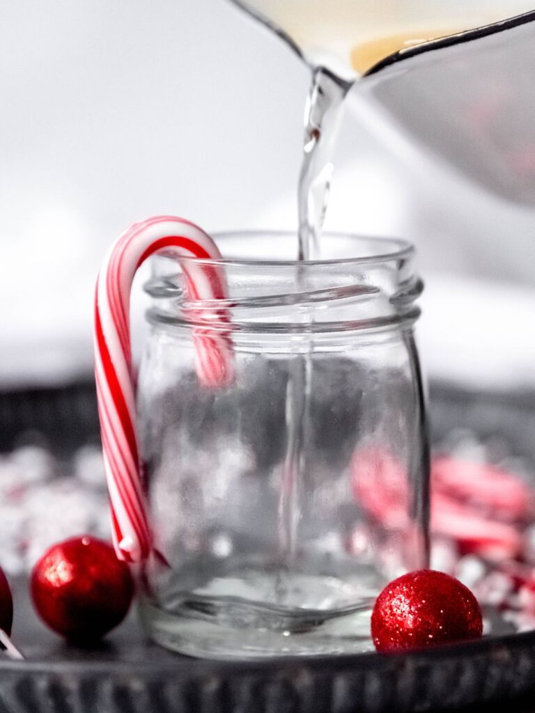 action shot of decanting the homemade peppermint syrup into a small glass jar with a mini candy cane hanging off the side.