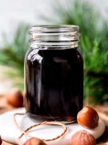 side-on shot of a jar of copycat starbucks hazelnut syrup in a glass jar in front of a branch of an evergreen tree with whole hazelnuts in the foreground.