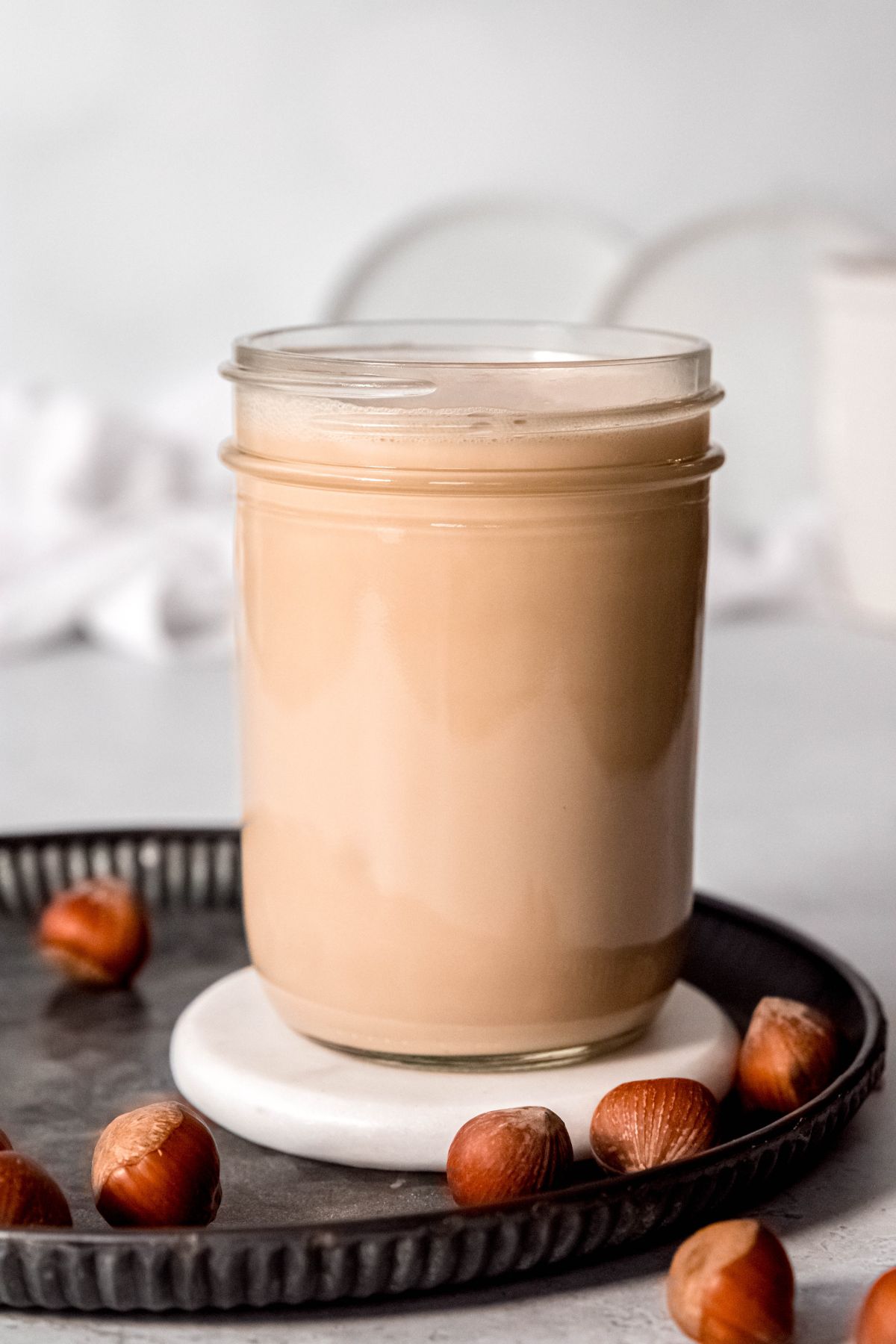 A side shot of a glass jar of hazelnut homemade coffee creamer on a metal tray with hazelnuts.
