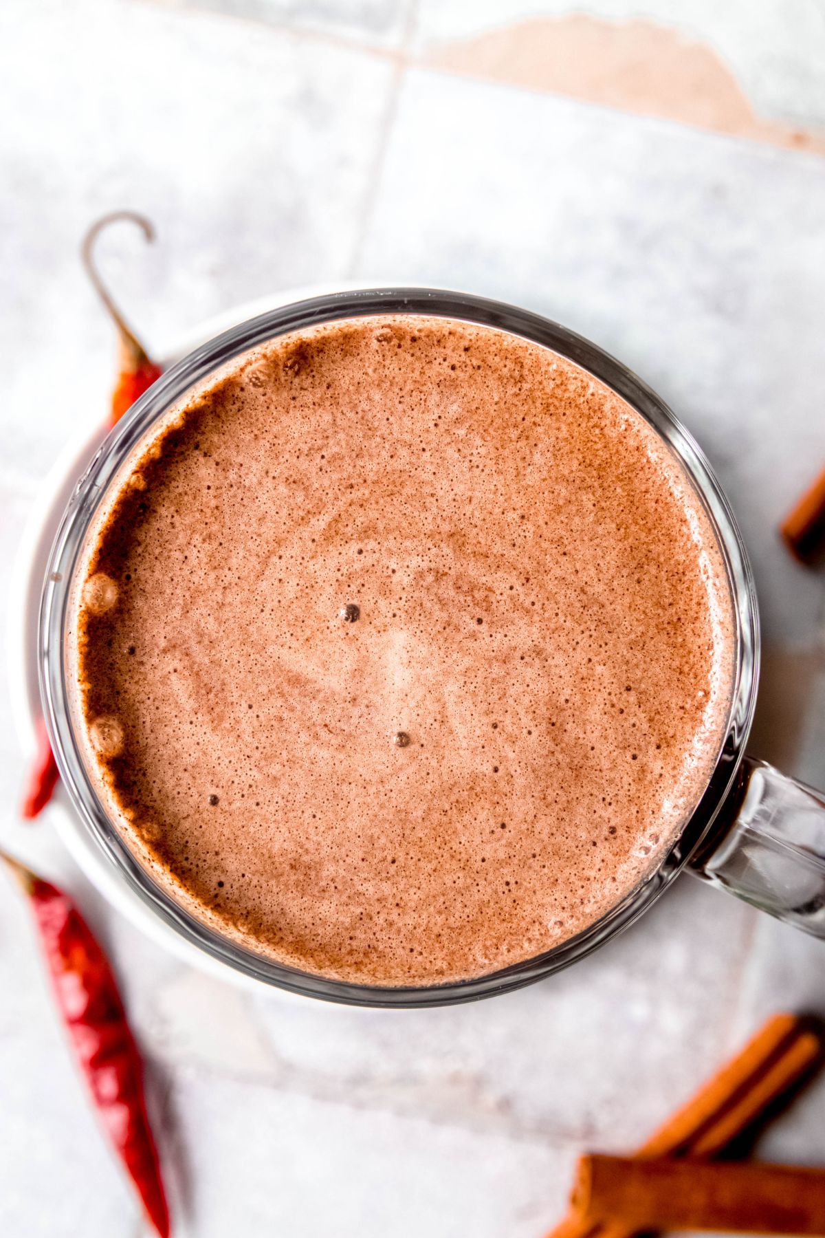 An overhead shot of a glass mug of Mexican mochas with dried peppers and cinnamon sticks in the background.
