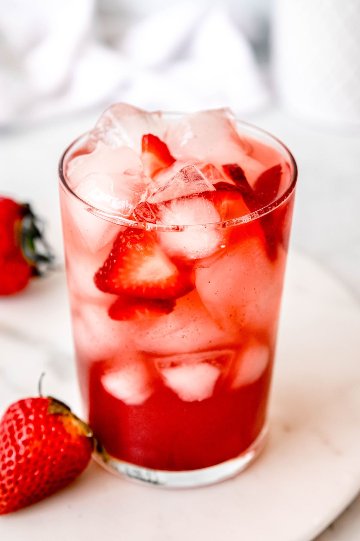 clear glass of a DIY Starbucks strawberry acai refresher on a white marble tray with whole strawberries scattered around. 