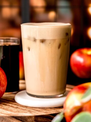 A side shot of a glass of apple cream chai latte on a coaster with apples.