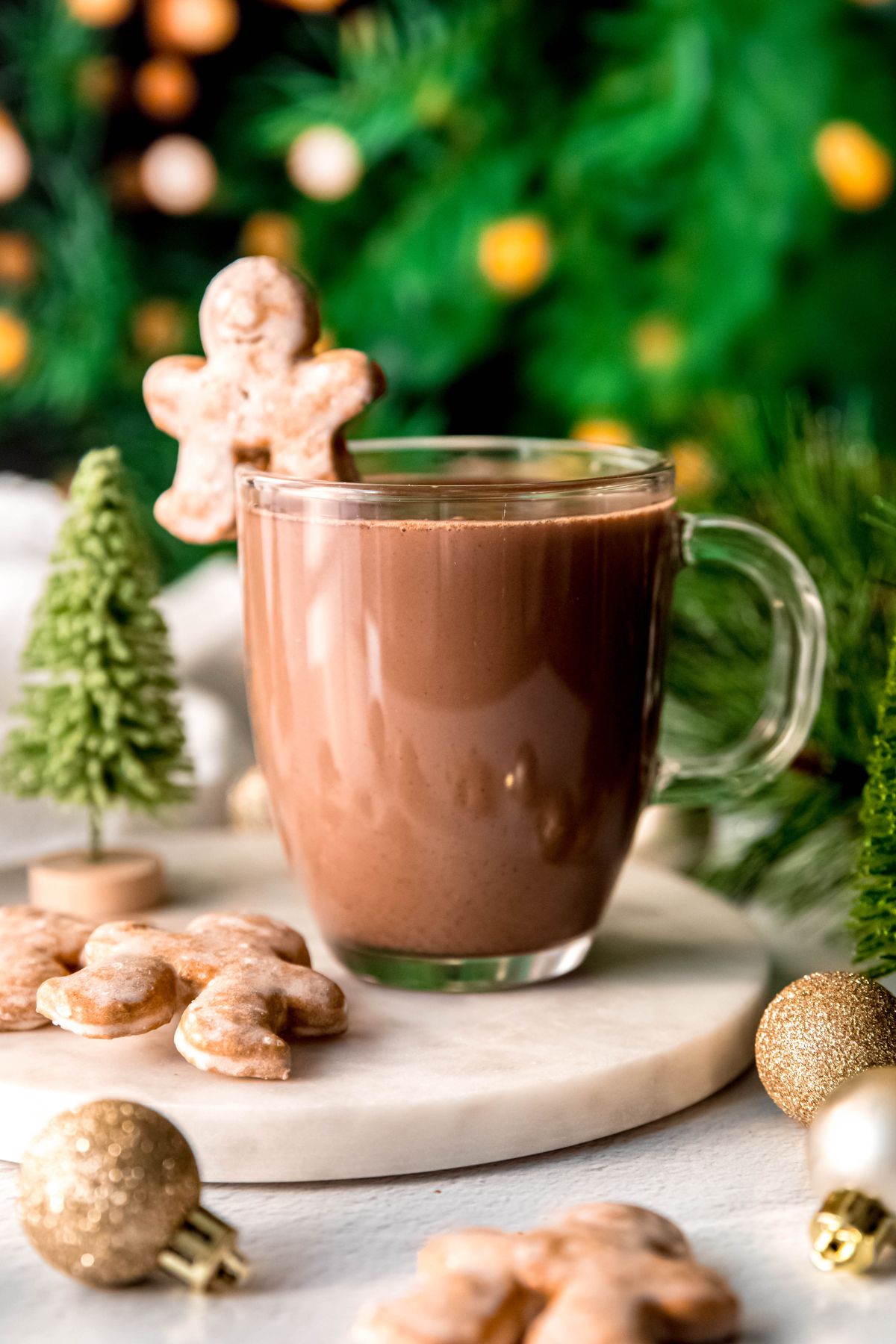 A close-up shot of a glass mug of gingerbread hot chocolate on the counter with gingerbread cookies, baubles, and evergreen branches. 