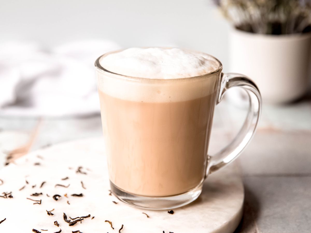 horizontally framed hero shot of a homemade london fog latte in a clear mug on a white table with dried tea leaves and a vase with dried lavender. 