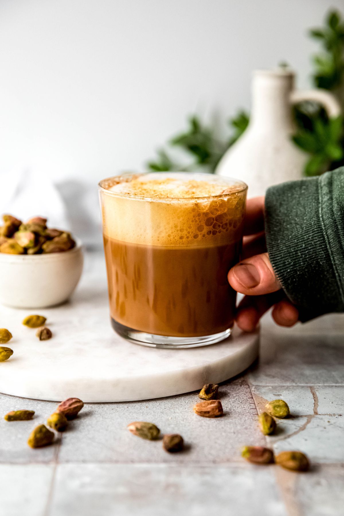 hand clad in a green sweater to the knuckles grabbing the clear glass of pistachio cortado from the table with the pistachios scattered around. 