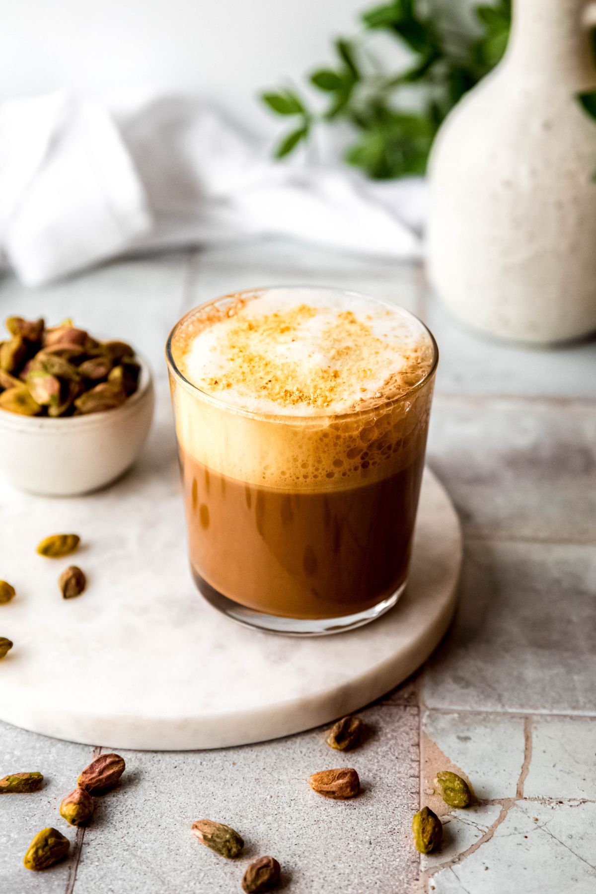 45 degree angle of a homemade starbucks pistachio oat milk cortado showing the crushed biscoff topping on the foam; its on a table with a pinch bowl of shelled pistachios with some spilling out of the bowl. 