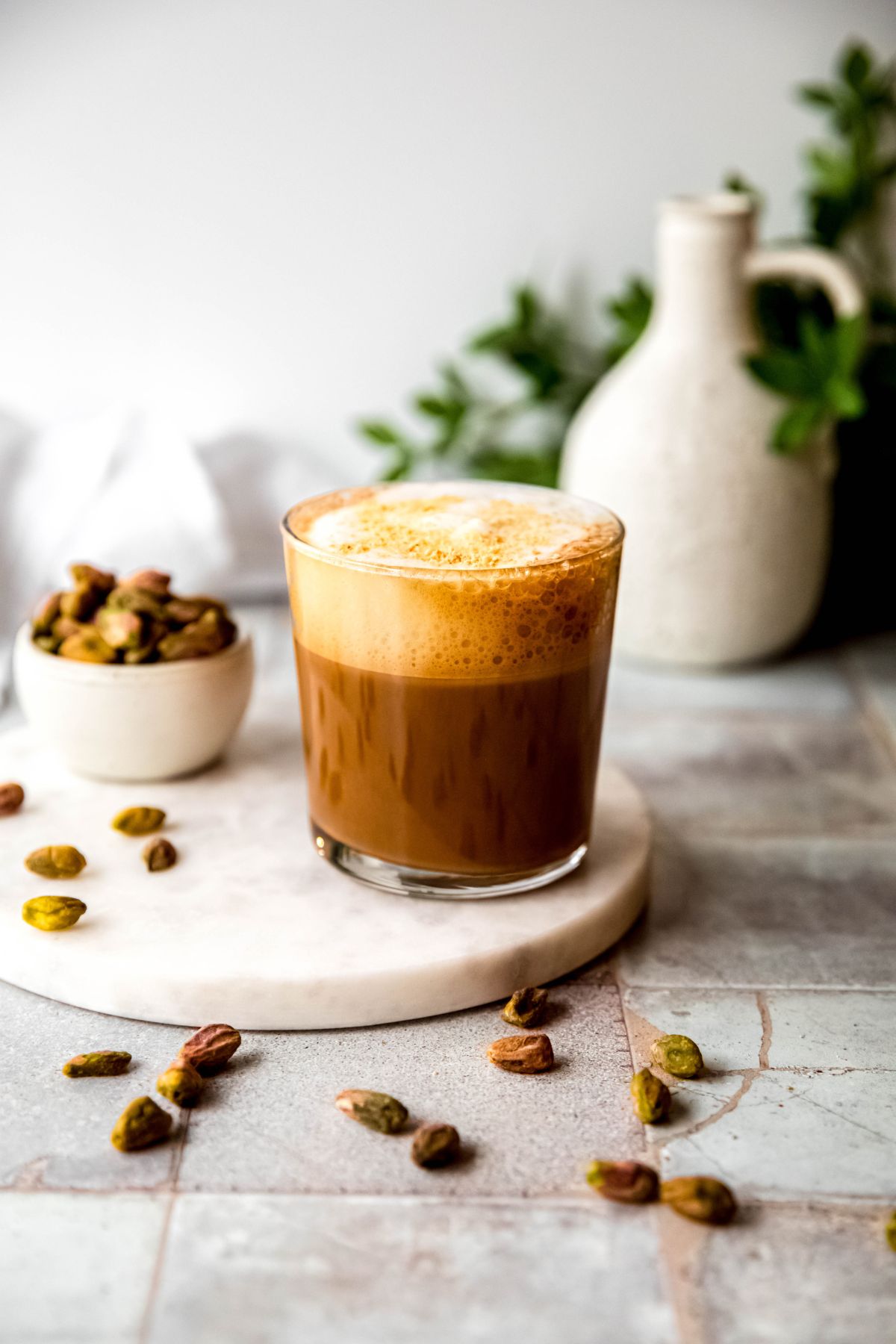 homemade pistachio oat milk cortado on a white marble round on a grey slate tile table with shelled pistachios scattered around and a green houseplant blurred out in the back. 