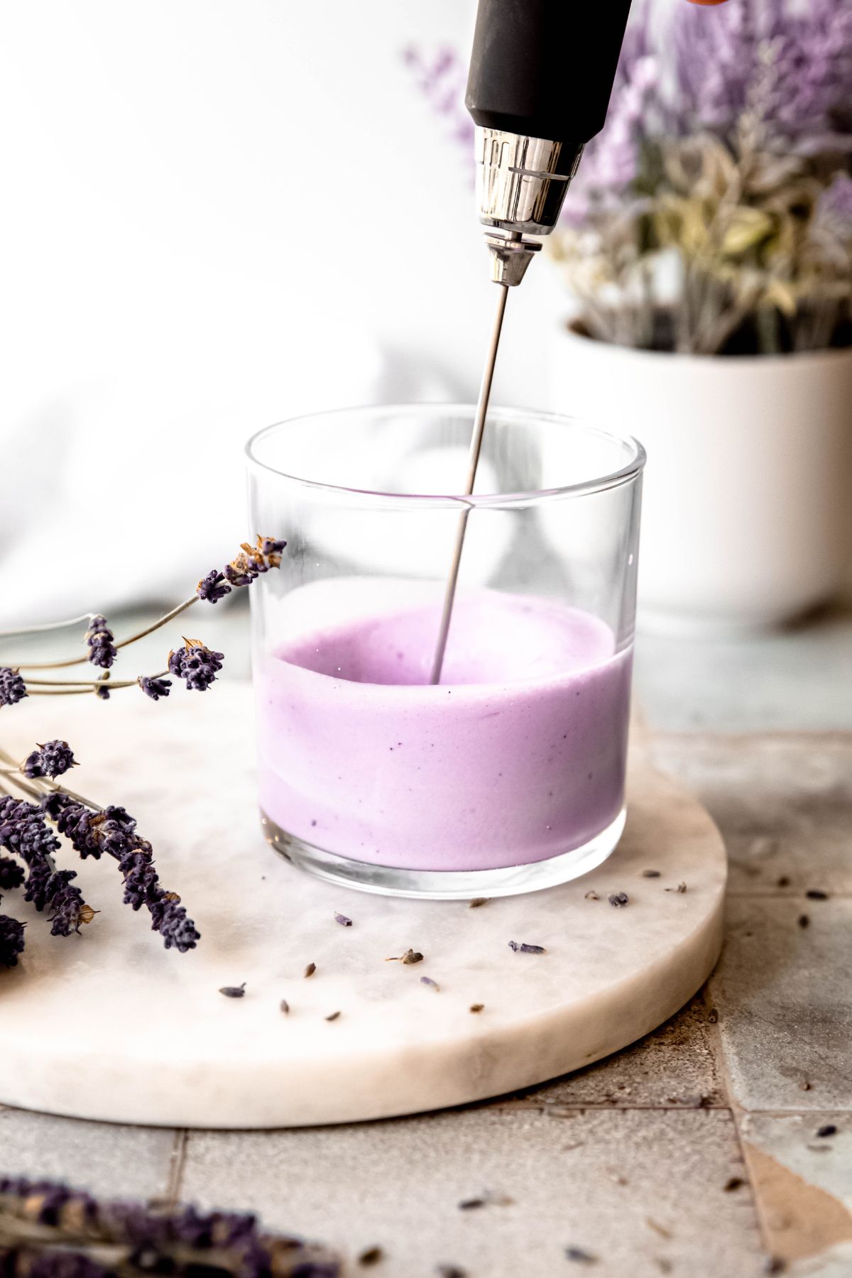 action shot of a milk frother frothing homemade lavender cold foam in a clear glass on a white table with dried lavender sprigs. 