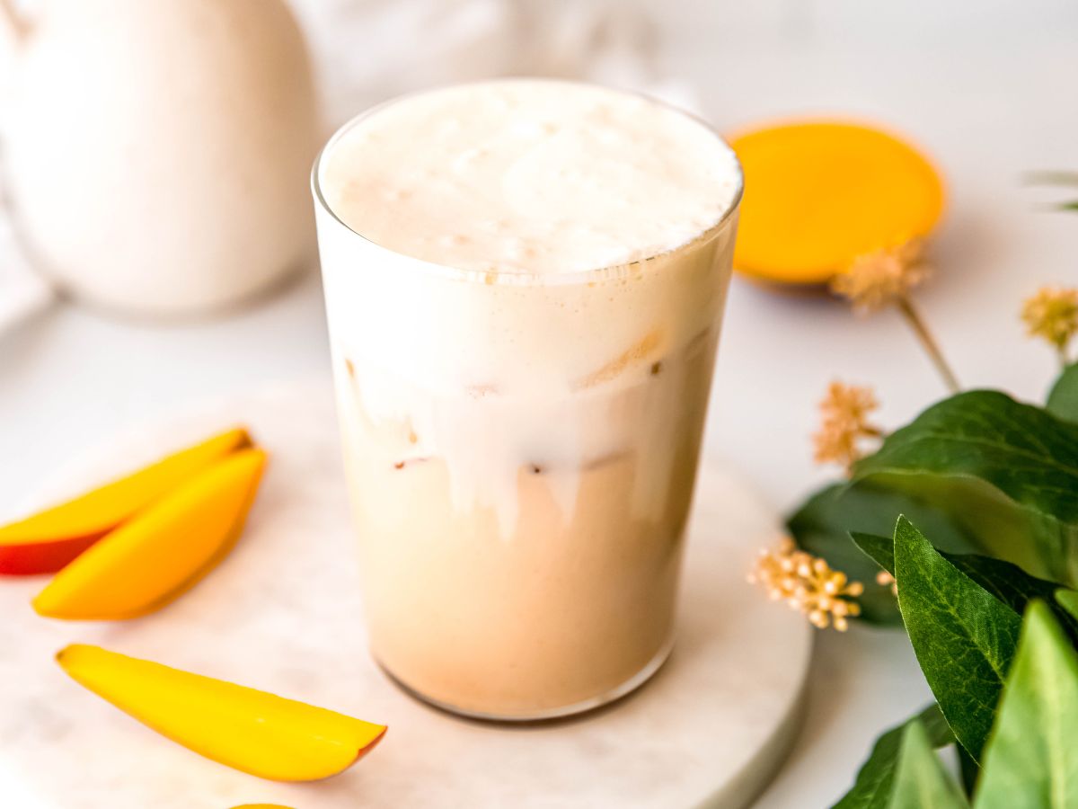 horizontally framed hero shot of a DIY iced mango cream chai tea latte on a white table with fresh mango slices and a green houseplant with yellow buds.