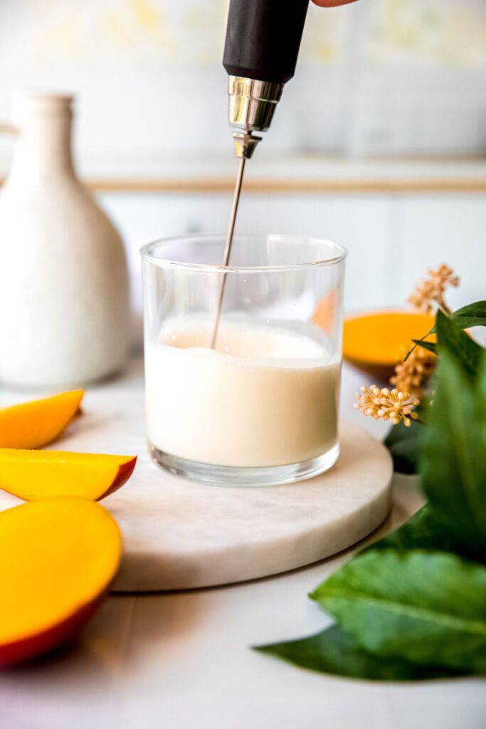 action shot of a milk frother making mango cream cold foam in a small rocks glass. 