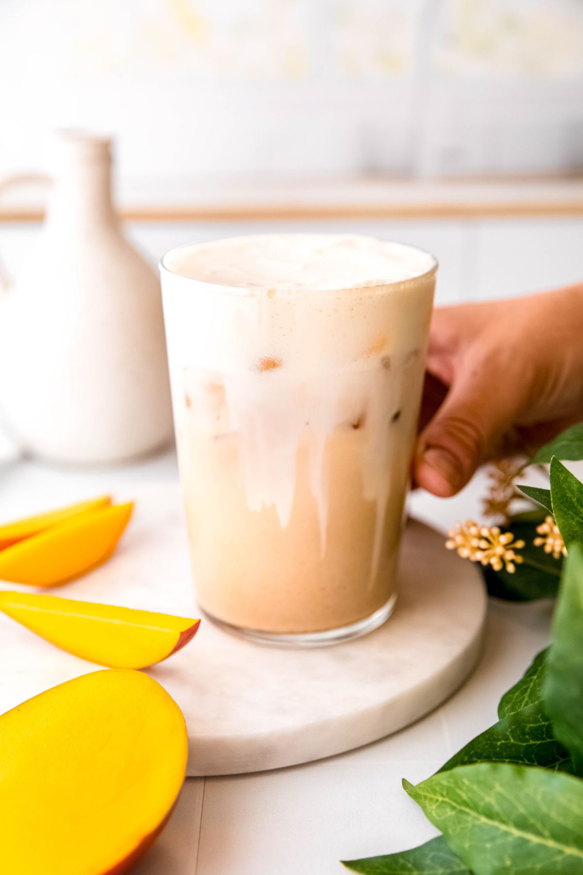 hand grabbing a pint glass filled with iced mango chai with mango cream cold foam from a white table with fresh mango slices and a green houseplant. 