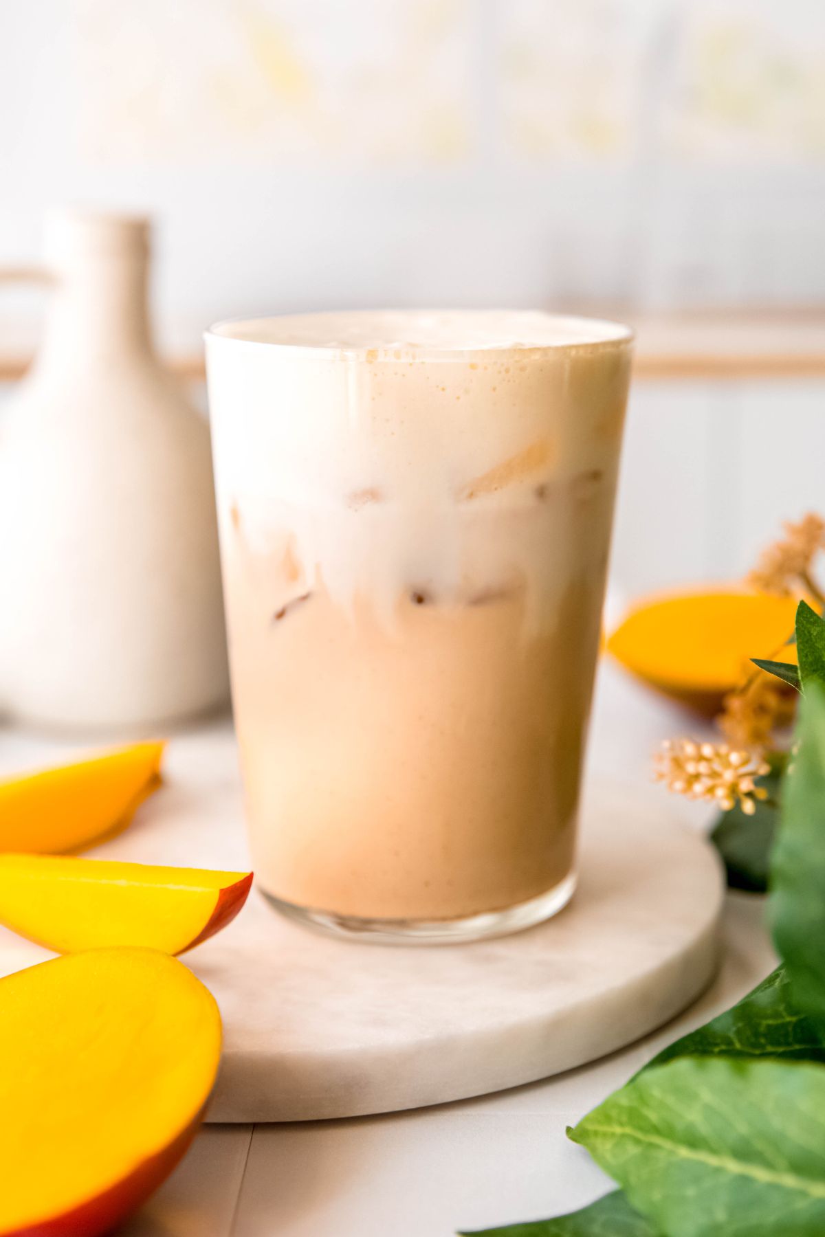 homemade iced mango cream chai latte in a clear pint glass on a white table with fresh mango slices. 