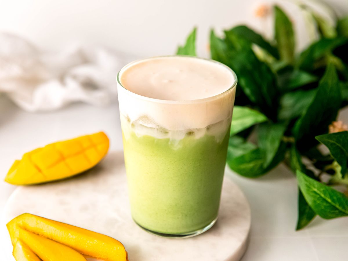horizontally framed hero shot of a DIY starbucks copycat iced mango matcha with mango cream cold foam on a white table with fresh mango slices and a green houseplant with yellow blooms. 