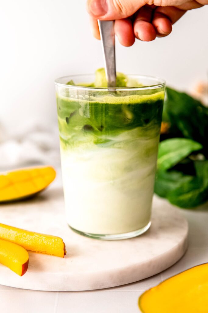 action shot of a hand stirring a silver spoon to mix the dark green matcha layer on top with the mango milk mixture below. 