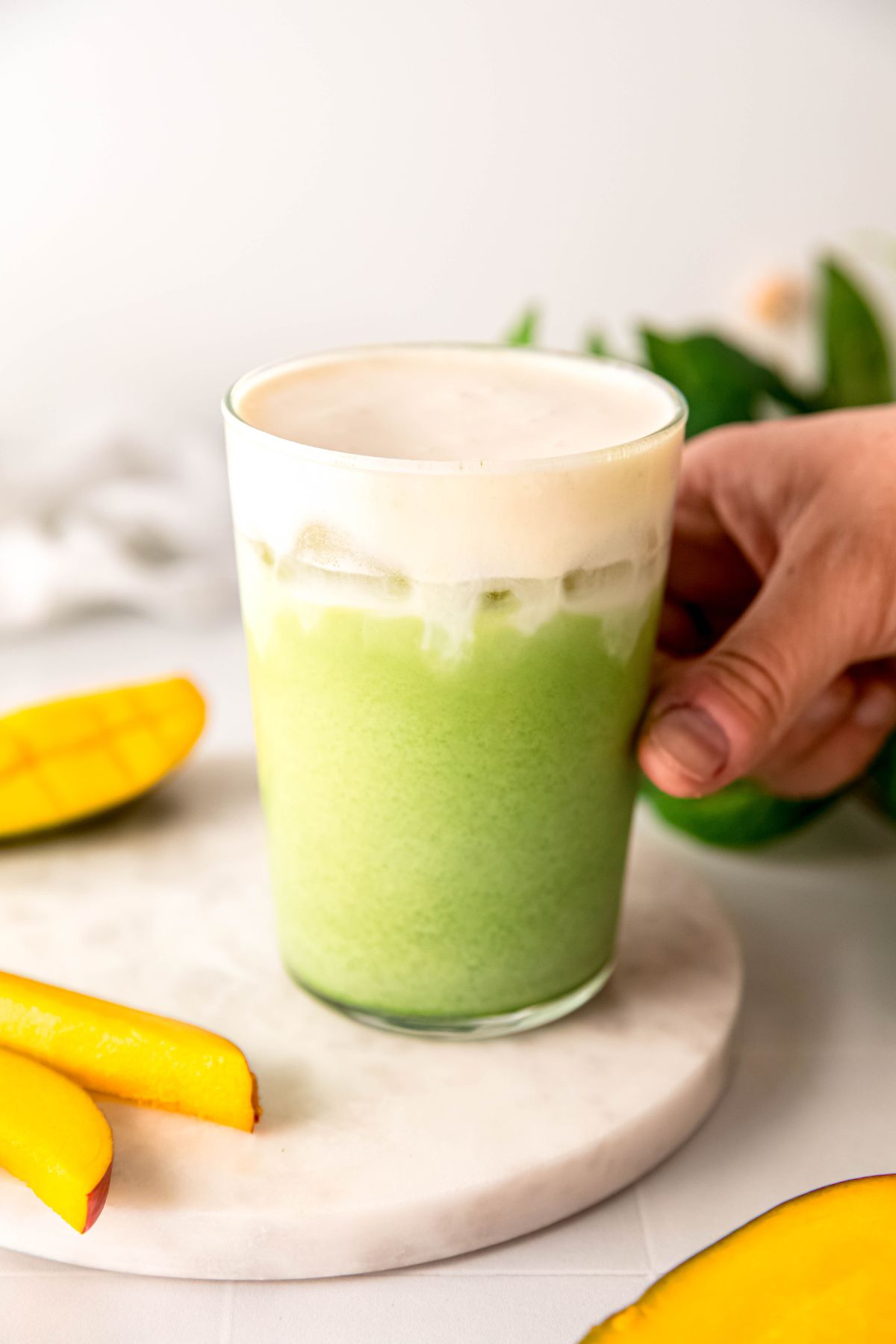 hand grabbing a clear pint glass with a DIY iced mango cream matcha from a white table with fresh mango slices. 