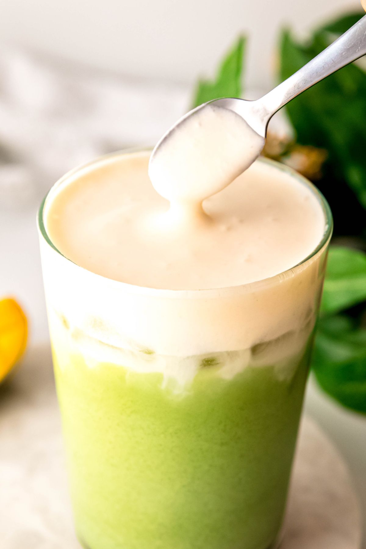 action shot of a silver spoon dolloping pale yellow mango cream cold foam on an iced matcha base. 