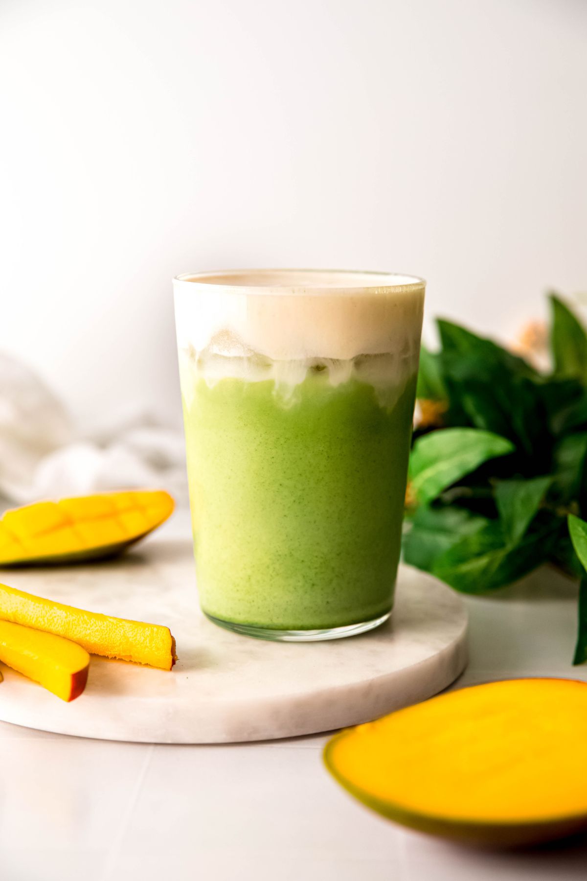 side on view of a pint glass filled with iced mango matcha latte topped with mango cream cold foam on a white table with fresh mango slices and a halved mango. 