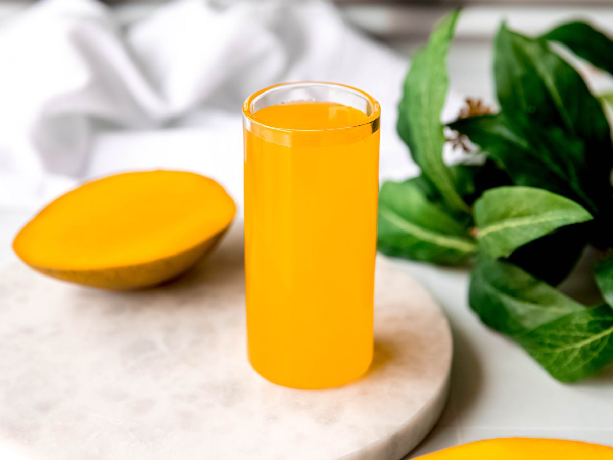 horizontally framed hero shot of fresh mango syrup on a white table with a halved mango and a green houseplant.