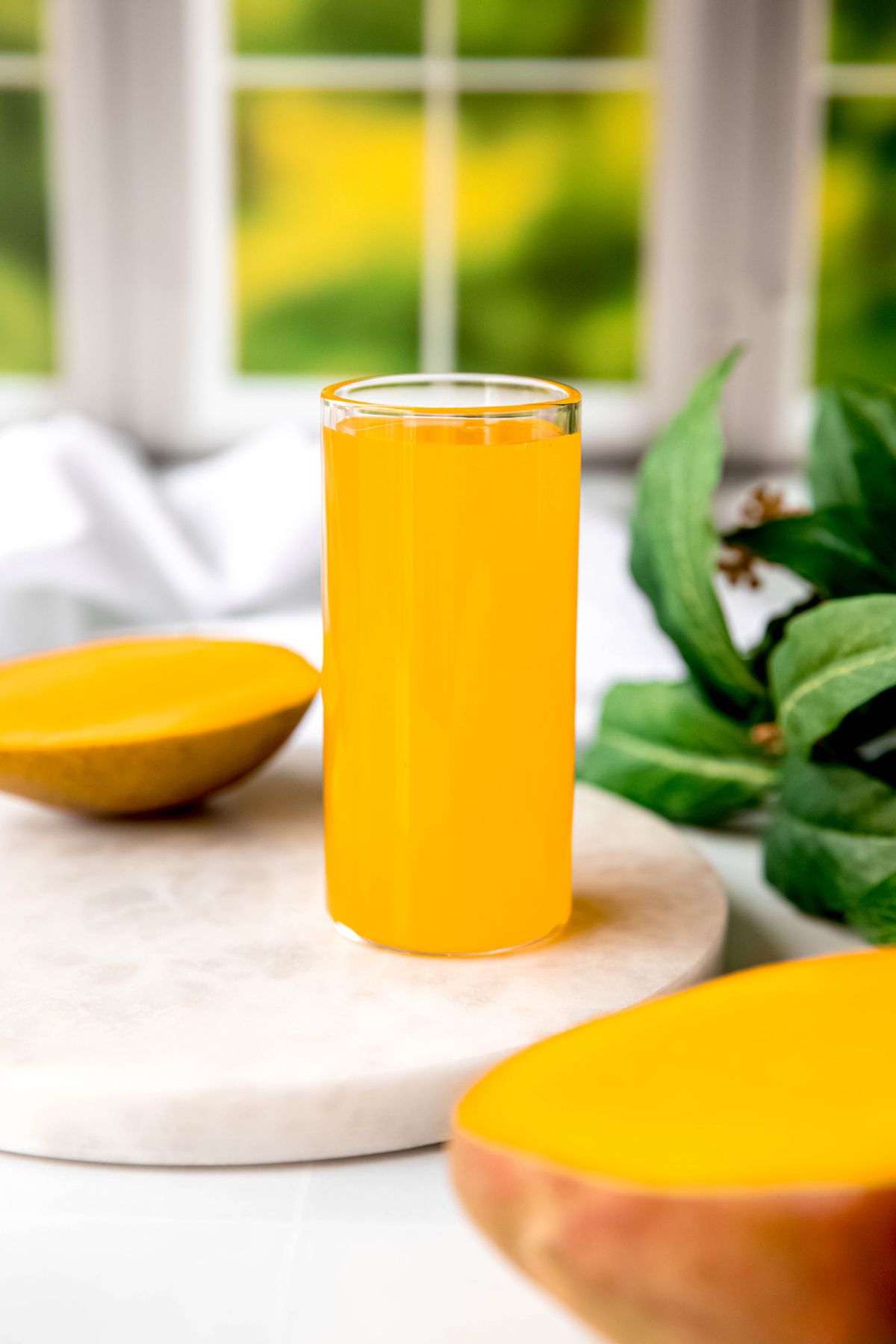 collins glass of homemade mango syrup on a white table in front of a window with green outdoors.