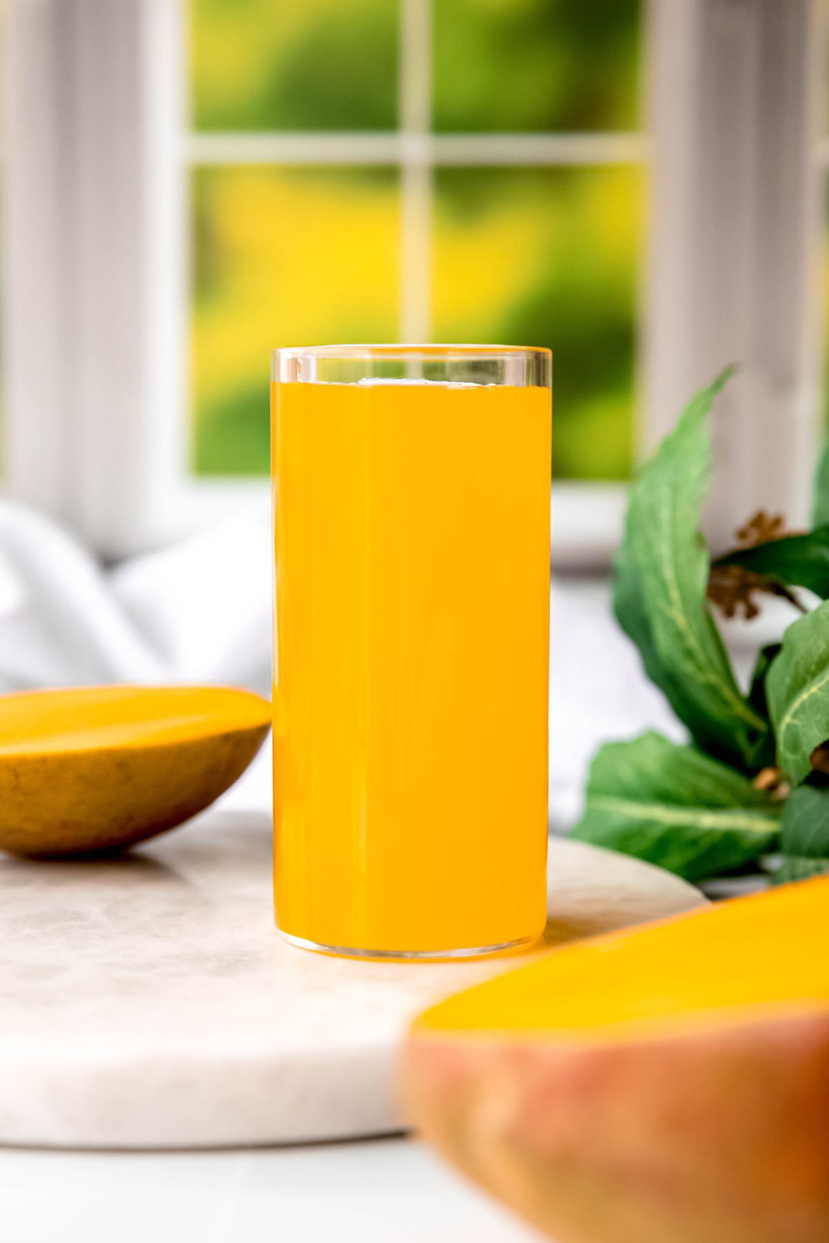 glass of homemade mango syrup for tea and drinks on a white table  with halved mango and a green houseplant in front of a window looking out on a green yard.