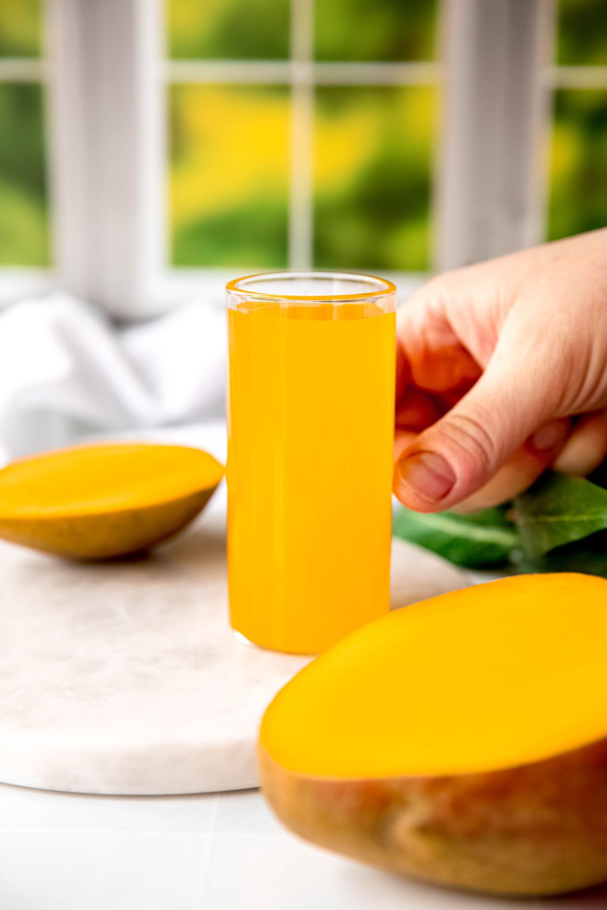 hand setting a collins glass of bright orange mango simple syrup down on a white table with a halved mango.