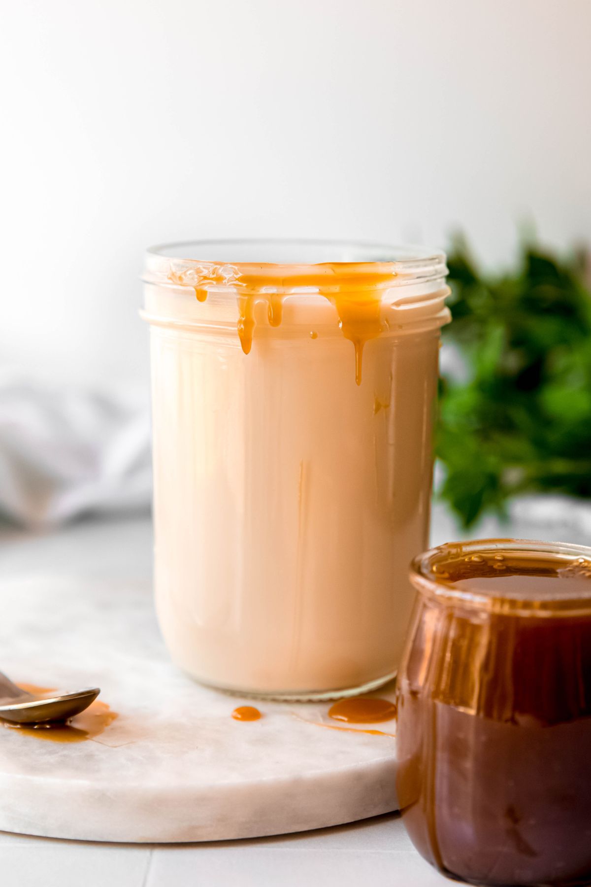 mason jar of homemade caramel macchiato coffee creamer on a white table with a jar of caramel sauce in the foreground and a dribble of caramel sauce running down the side of the mason jar filled with the creamer. 