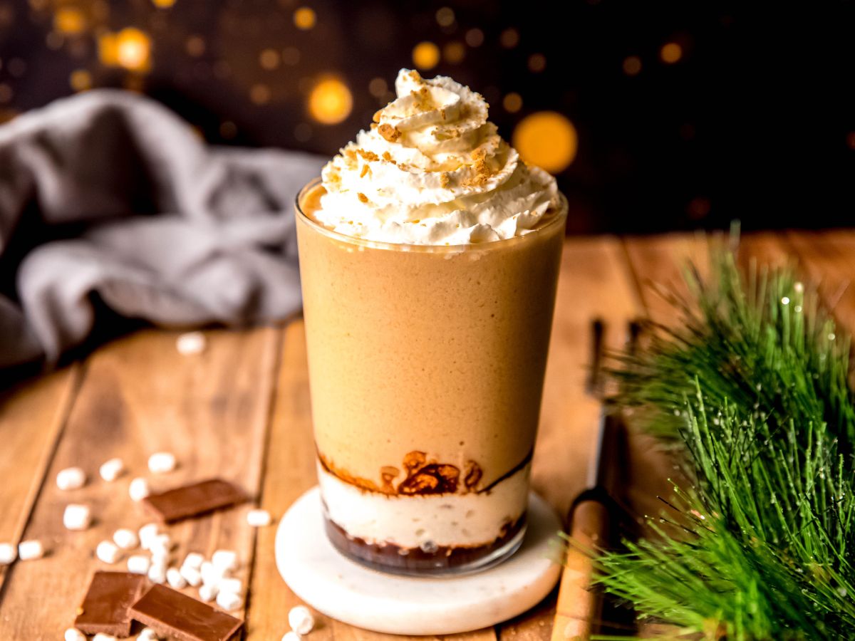 horizontally framed hero image of a homemade s'mores frappe in a clear pint glass on a rustic wooden table with marshmallows, chocolate bars, and a sprig of pine tree in front of a dark night sky with a fire. 