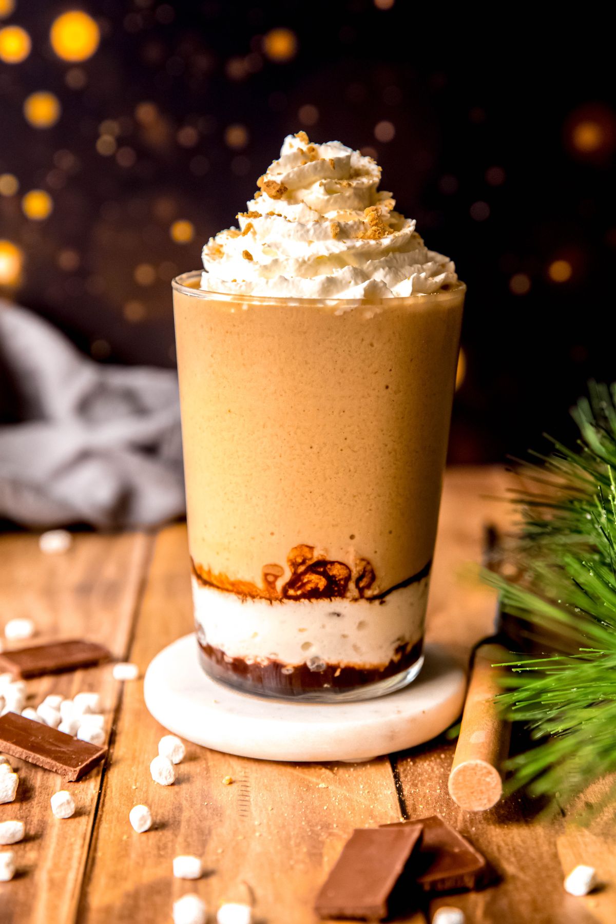 hero shot of a DIY Starbucks s'mores frappuccino on a wooden table with graham cracker crumbs, mini marshmallows, chocolate pieces, a green pine sprig, and a marshmallow roasting fork in front of a black sky with a campfire. 