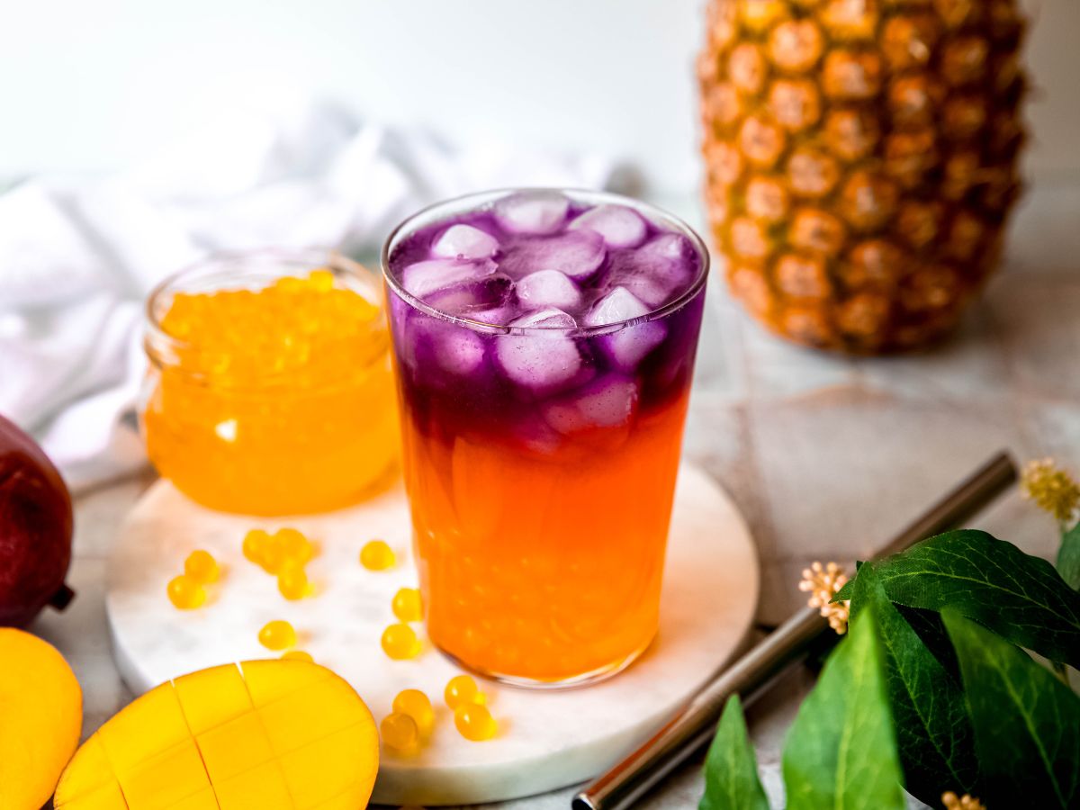 horizontally framed hero image of a homemade tropical butterfly refresher from starbucks on a white table with a whole and halved mango, a whole pineapple, a jar of mango boba with some pearls spilling over onto the table, a green houseplant with yellow buds, and a silver boba straw.