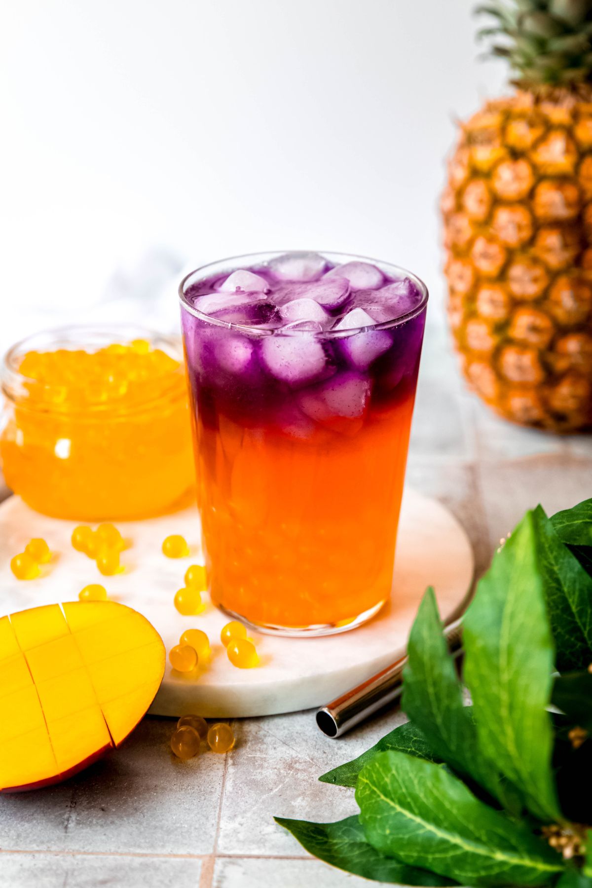 homemade tropical butterfly starbucks refresher in a clear pint glass on a white marble round on a grey slate table with a green houseplant and a halved mango in the foreground and a pineapple and a jar of mango popping boba in the background. 