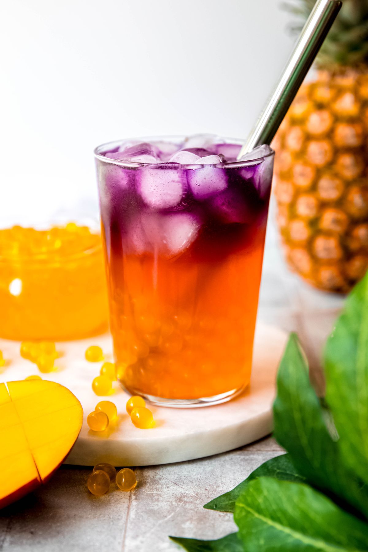 homemade tropical butterfly refresher in a clear pint glass on a white table with a whole pineapple and a jar of mango popping boba blurred in the background and a halved mango and green houseplant in the fore.