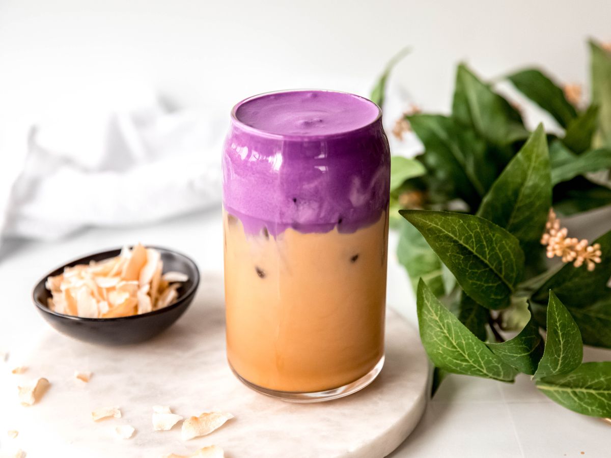 horizontally framed hero shot of a homemade iced coconut ube cream shaken espresso on white table with toasted coconut flakes scattered around with more in a small black pinch bowl and a green houseplant with yellow blooms. 