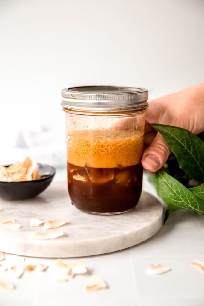 hand setting down a lidded mason jar with the coconut shaken espresso base on a white table. 