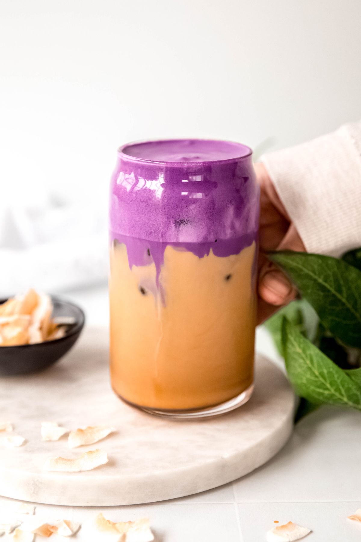 hand in a white sweater up to the knuckles grabbing a clear glass of iced ube coconut cream shaken espresso from a white table with toasted coconut flakes and a green houseplant. 
